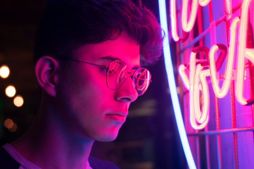 A young man with glasses illuminated by vibrant neon lights in an urban setting.